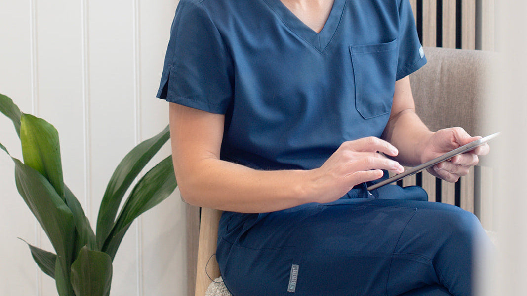 Person in blue scrubs sitting on a chair using a tablet.