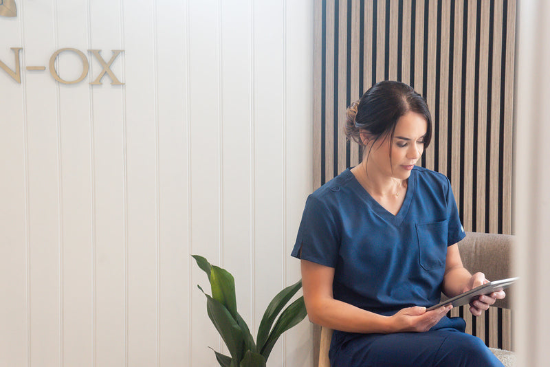 Woman in blue scrubs sitting and using a tablet in a room with a plant and wall decor.