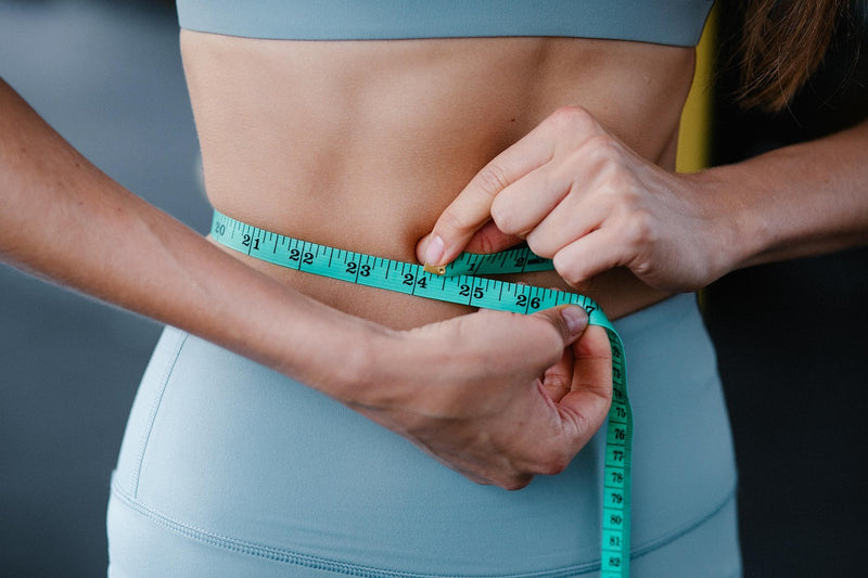 Person measuring waist with a green tape measure against a neutral background
